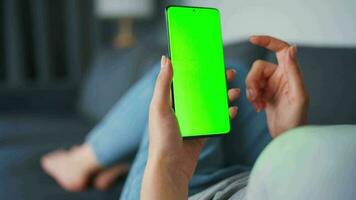 Woman at home lying on a couch and using smartphone with green mock-up screen in horizontal landscape mode. Girl browsing Internet, watching content, videos, blogs. POV. video