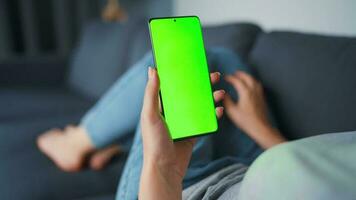 Woman at home lying on a couch and using smartphone with green mock-up screen in horizontal landscape mode. Girl browsing Internet, watching content, videos, blogs. POV. video