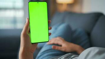 Woman at home lying on a couch and using smartphone with green mock-up screen in horizontal landscape mode. Girl browsing Internet, watching content, videos, blogs. POV. video