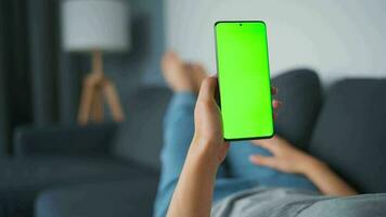 Woman at home lying on a couch and using smartphone with green mock-up screen in horizontal landscape mode. Girl browsing Internet, watching content, videos, blogs. POV. video