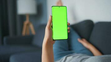 Woman at home lying on a couch and using smartphone with green mock-up screen in horizontal landscape mode. Girl browsing Internet, watching content, videos, blogs. POV. video