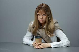 enojado infeliz bonito joven rubia estudiante dama persona de libre dedicación en calentar suéter con para llevar café sentado a lugar de trabajo en terminado gris moderno hogar oficina. café descanso amante concepto. Copiar espacio. frio oferta foto