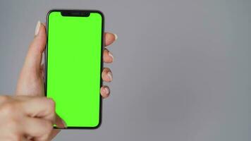 hembra manos utilizando un teléfono inteligente con un verde pantalla en un gris antecedentes de cerca. croma llave video