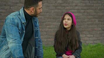 Dad and daughter are sitting and talking against the backdrop of grass and a brick wall. Concept of support and happy video