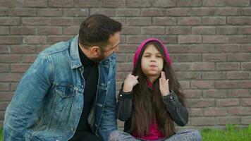 Dad and daughter are sitting and talking against the backdrop of grass and a brick wall. Concept of mutual understanding, support and happy family video