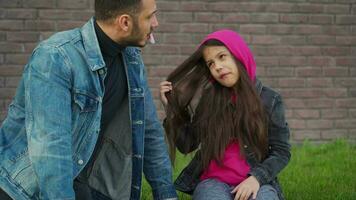 Dad and daughter are sitting and talking against the backdrop of grass and a brick wall. Concept of support and happy video