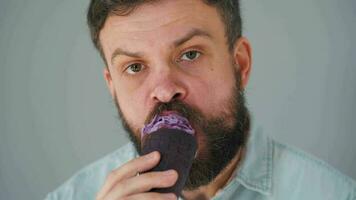 Nahansicht von ein bärtig Mann Essen lila Eis Sahne im ein schwarz Waffel Tasse auf ein grau Mauer Hintergrund video