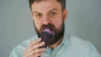 Nahansicht von ein bärtig Mann Essen lila Eis Sahne im ein schwarz Waffel Tasse auf ein grau Mauer Hintergrund video