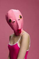 A woman wearing a pink fish head mask on Halloween stands and looks at the camera against a pink background. Conceptual art photo in a crazy way