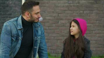 Dad and daughter are sitting and talking against the backdrop of grass and a brick wall. Concept of support and happy video