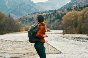 woman in sweater with a backpack rest in the mountains near the river in autumn in nature and tall trees model photo