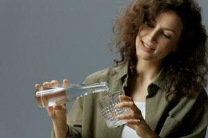 Happy smiling curly beautiful woman in casual khaki green shirt pours water from bottle in glass posing isolated on over gray blue background. Healthy lifestyle. Water is Life concept. Free place photo