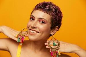 Portrait of a young woman with a short haircut and colored hair smiling and showing her tongue at the camera on an orange background with earrings accessories in the studio photo