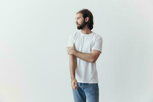 A man with a beard and long hair in a white T-shirt and blue jeans stands against a white wall leaning full-length on it, relaxed style photo