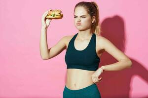 sportive woman with hamburger in hands eating food pink background fast food photo