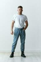 Full body shot of handsome serious tanned man guy in basic t-shirt holds hand on jeans posing on white background. Fashion Style New Collection Offer. Copy space for ad. Modeling snapshots photo