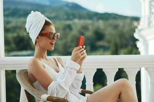 hermosa mujer en un blanco camisa se sienta en el balcón con un teléfono perezoso Mañana foto
