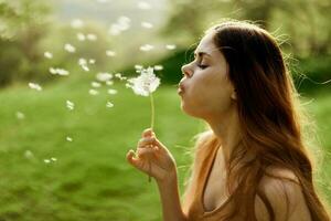 Woman with a dandelion flower in her hands smiling and blowing on it against a background of summer greenery and sunlight photo