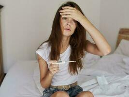 a woman holds her hand on her forehead while sitting on the bed with a thermometer in her hands photo