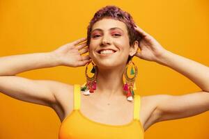 Portrait of a young woman with a short haircut and colored hair smiling and showing her tongue at the camera on an orange background with earrings accessories in the studio photo