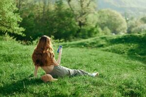Top view of a woman in an orange top and green pants sitting on the summer green grass with her back to the camera with her phone, a young freelance student's concept of work and leisure photo