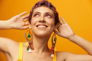 Portrait of a young woman with a short haircut and colored hair smiling and showing her tongue at the camera on an orange background with earrings accessories in the studio photo