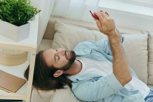 un hombre con un barba mentiras en el sofá durante el día a hogar y mira a su teléfono relajante en su día apagado, un hombre juego en el valores mercado en línea en su teléfono foto