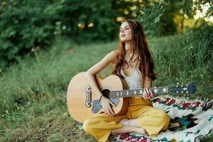 Girl hippie woman playing guitar in eco-friendly clothes sitting on the ground outside in nature in the fall watching the sunset photo