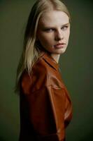 Closeup studio portrait of blonde with expressive glance blue eyes showy makeup looking over her shoulder at camera. Beautiful young woman in trendy leather brown dress posing isolated on green wall photo