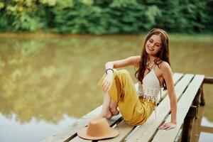 A young hippie woman sits on a lake bridge wearing stylish eco clothes and smiling photo