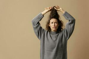 Problematic unruly damaged hair concept. Confused curly beautiful woman in gray casual sweater with hairbrush comb pulls tangled hair up posing isolated on over beige pastel background. Copy space photo