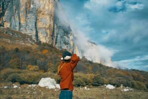 mujer en un suéter y pantalones con elevado arriba brazos viajes en el montañas en naturaleza paisaje foto