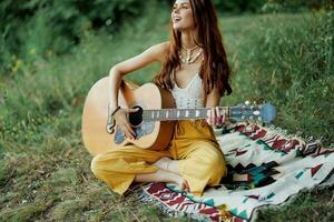 Young female hippie artist plays guitar and sings songs in eco-friendly clothing sitting on the ground outside in nature in the autumn looking out at the sunset photo