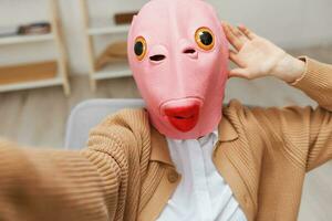 Funny blonde lady in pink fish mask warm sweater doing selfie use phone sitting in armchair at modern home interior. Pause from work, take a break, social media in free time concept. Wide angle photo