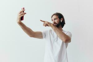 A male blogger videotaping himself on his phone and chatting with people online with a smile in a white T-shirt against a white wall photo
