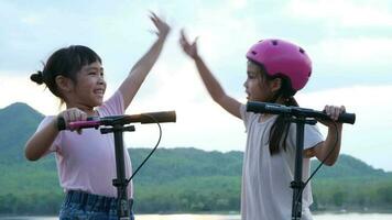 Two cute little girls smiling and posing together in summer garden. Happy kid riding kick scooter in the park. Healthy sports and outdoor activities for children. video