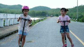 Two cute little girls riding kick scooters on outdoor road in summer park. Healthy sports and outdoor activities for school children in the summer. video