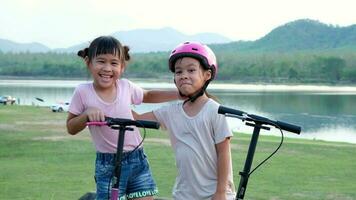 Two cute little girls smiling and posing together in summer garden. Happy kid riding kick scooter in the park. Healthy sports and outdoor activities for children. video