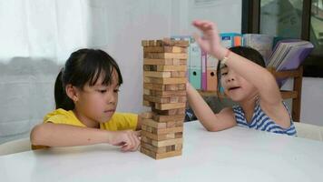 söt asiatisk syskon har roligt spelar jenga tillsammans. två barn spelar jenga styrelse spel på tabell i rum på Hem. trä- pussel är spel den där öka intelligens för barn. video