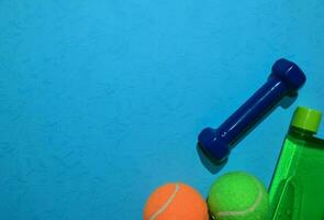 blue dumbbell, Green and orange tennis ball, and bottle indicating workout plan on blue background. Healthy and fitness concept. photo