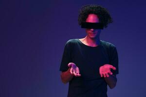 Excited stylish tanned curly man in black t-shirt eyewear holds invisible object posing isolated on color pink blue background. Cool fashion offer. Virtual Reality New Collection concept. Copy space photo