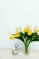 Easter eggs in a glass and yellow tulips in a vase on a light background photo