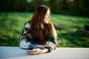 Girl with phone in hand sitting in nature in the park at the table smiling beautifully and looking at the camera with her red hair lit by the sunset sunlight of summer photo