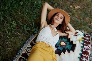 un mujer en Respetuoso del medio ambiente ropa en un hippie Mira es acostado en un de colores tartán sonriente y mirando a un otoño puesta de sol en naturaleza en el parque. estilo de vida en un viaje de armonía con el mundo foto