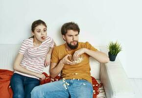 un hombre y un mujer en un brillante habitación sentar en el sofá con palomitas de maiz en un plato emociones amigos verde flor en un maceta foto