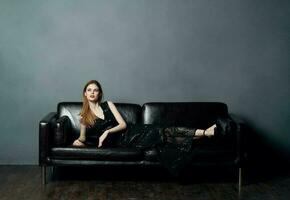 An elegant lady in a black dress lies on a leather sofa indoors in full growth photo