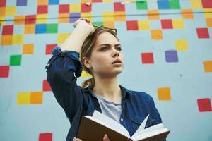 A student in a shirt and glasses holds a book in her hands on the street near the graffiti on the wall photo