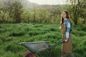 A young woman rolls a garden cart with soil for planting in her green nature garden and smiles photo