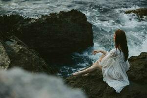 Barefoot Woman in White Dress with Wet Hair Sits on a Cliff photo