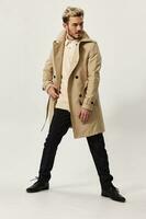 fashionable man in a beige coat looks to the side on a light background and dark trousers model in full growth photo